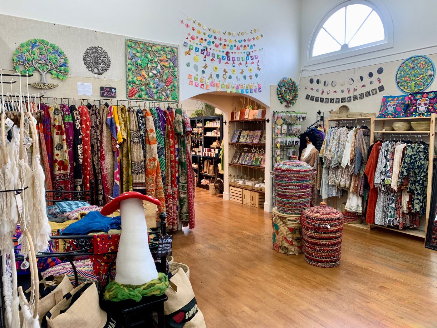Racks of printed clothing and scarves under an arched window, with a garland of felt hearts and woven baskets on the wood floor